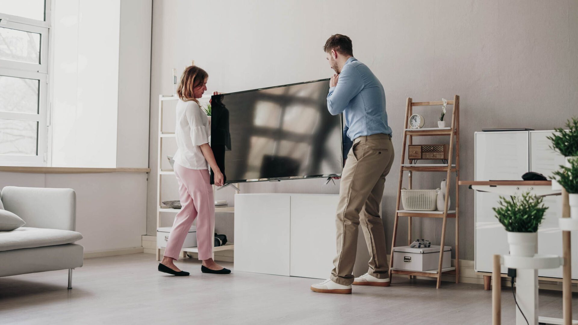 En kvinna och en man lyfter en platt-TV tillsammans i ett ljust, modernt vardagsrum med ljusa möbler, hyllor och växter.