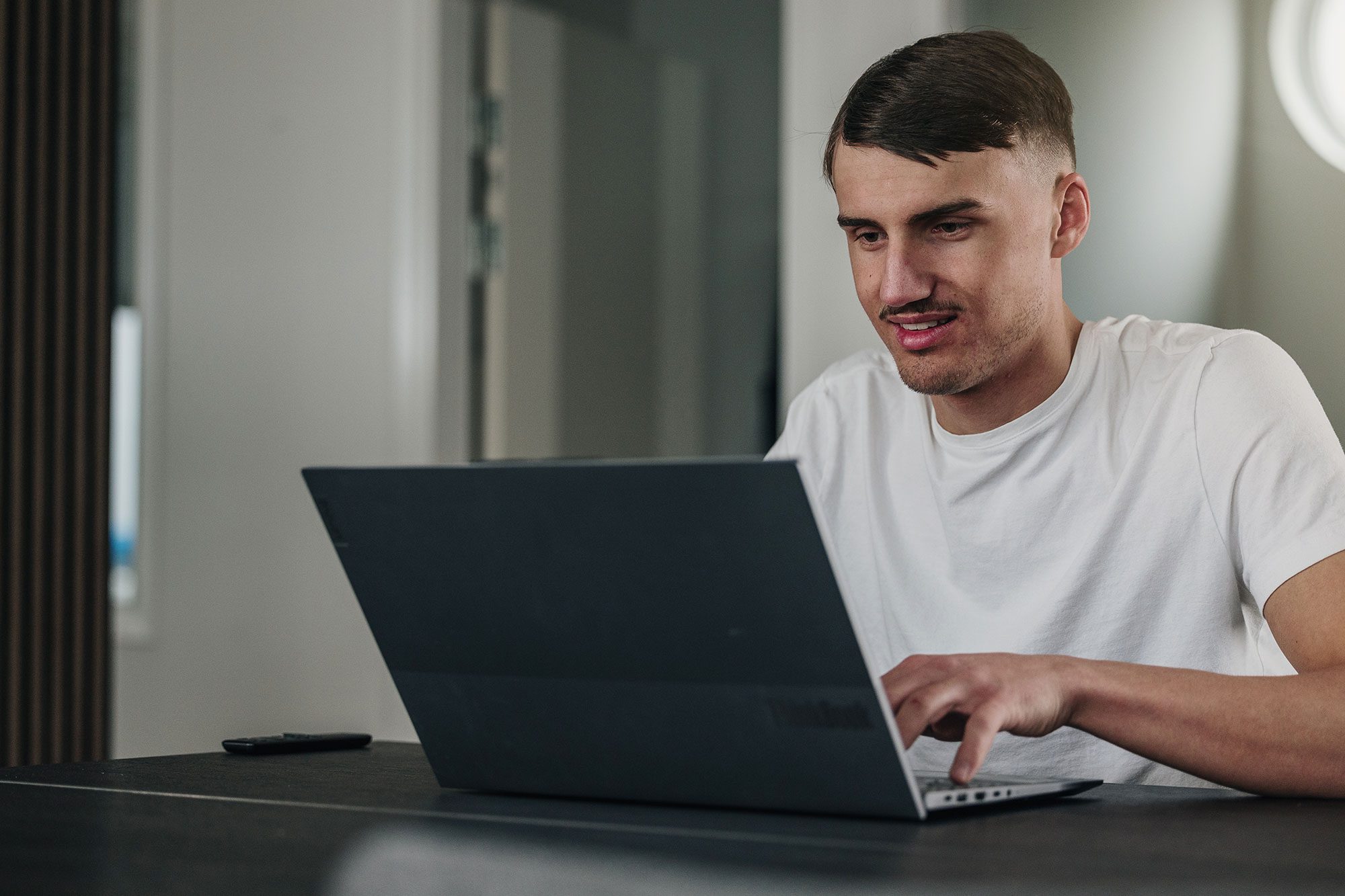 En person i vit t-shirt sitter vid ett bord och använder en svart laptop. En smartphone ligger på bordet och i bakgrunden syns en dörr och en rund lampa på väggen.