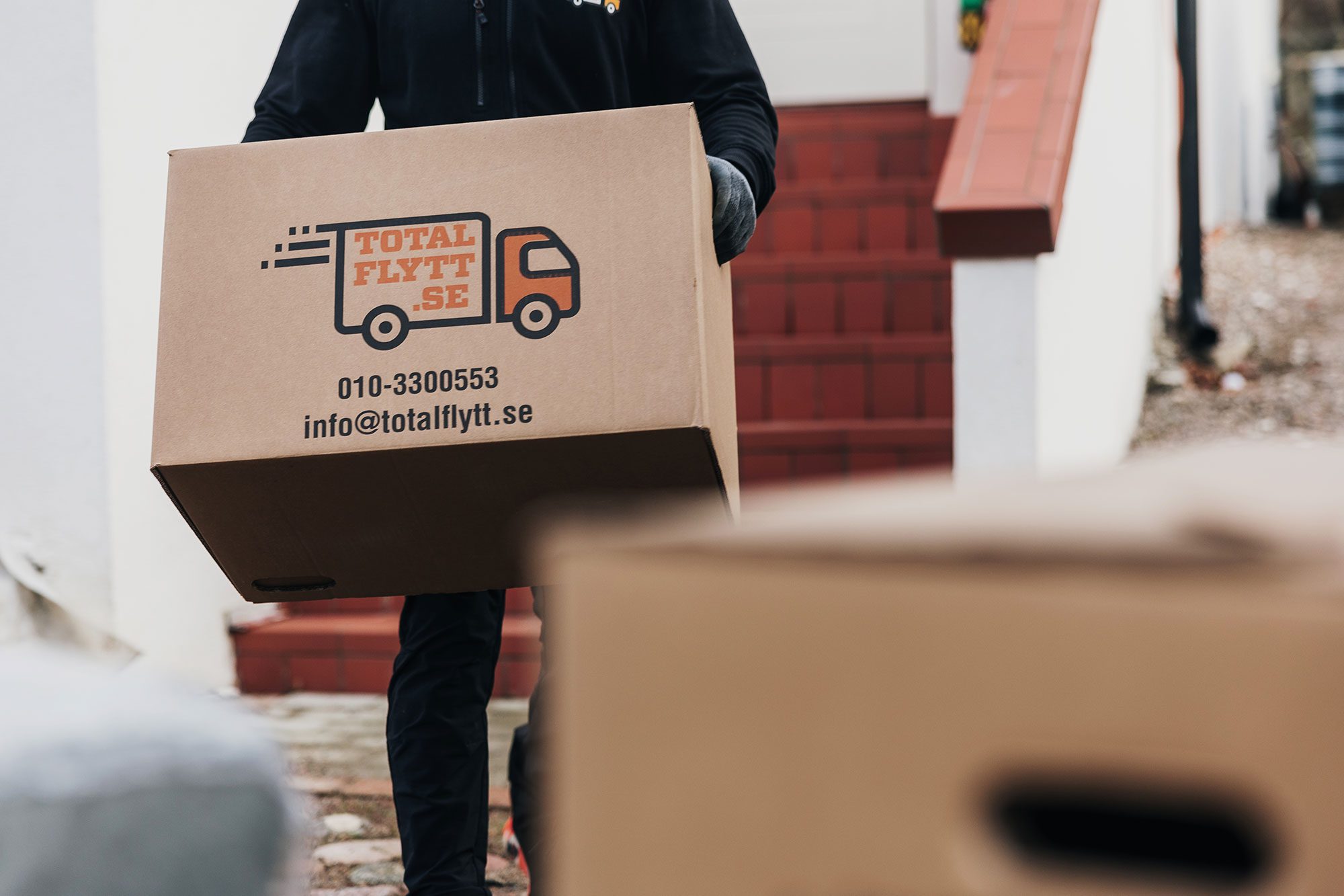 En person i mörka kläder bär en stor kartong märkt "Total Flytt SE" med kontaktuppgifter tryckta på den. En annan låda står i förgrunden. Röda trappor och en del av en byggnad syns i bakgrunden.