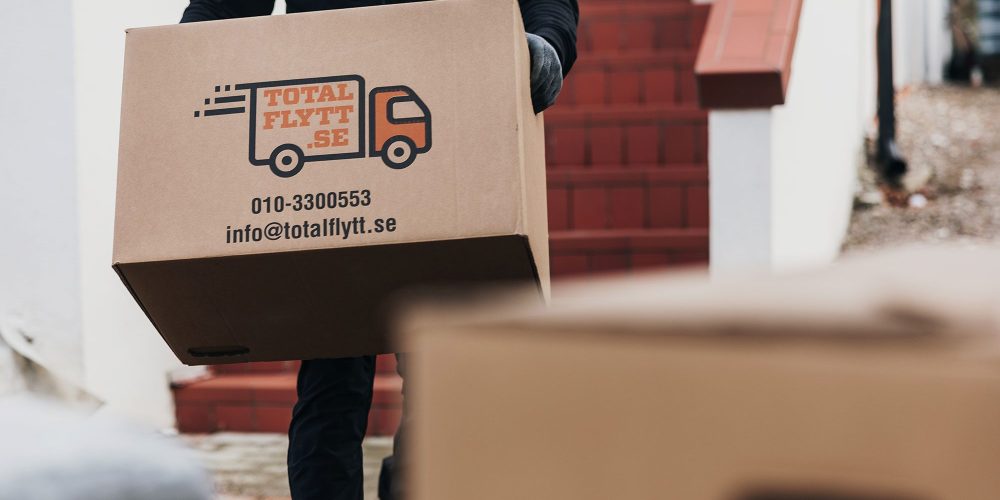 En person i mörka kläder bär en stor kartong märkt "Total Flytt SE" med kontaktuppgifter tryckta på den. En annan låda står i förgrunden. Röda trappor och en del av en byggnad syns i bakgrunden.