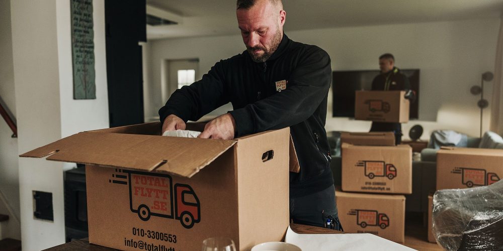 En man packar saker i en stor flyttlåda av kartong märkt "Total Flytt" i ett vardagsrum. En annan person står i bakgrunden, och uppackade glas och tallrikar står på bordet.