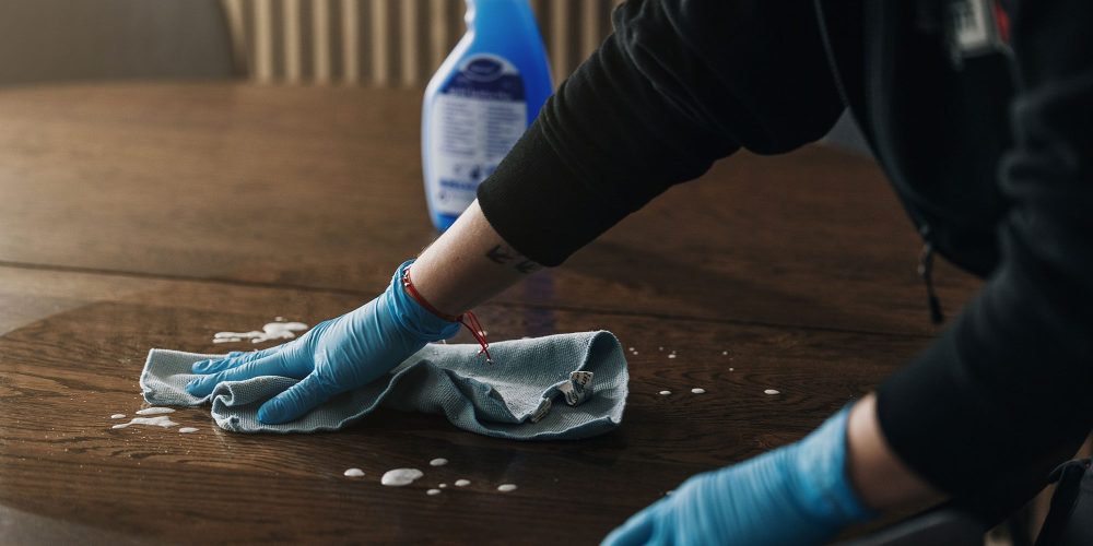 En person med blå handskar torkar av ett träbord med en trasa och rengör flytande rengöringsmedel. En sprayflaska med rengöringslösning står på bordet intill.